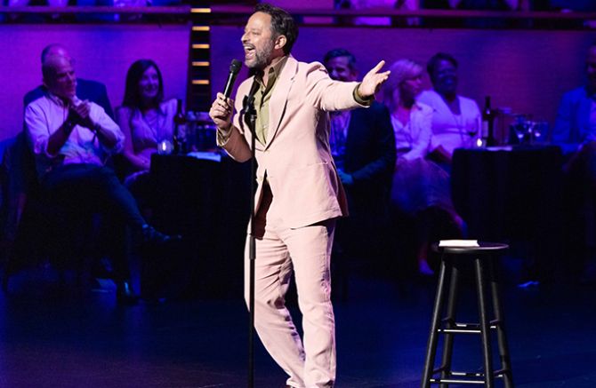 Comedian in a pink suit performs on stage, audience laughing.