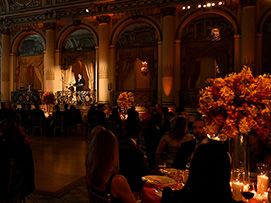 Ornate ballroom with people seated, warm lighting, and floral centerpieces.