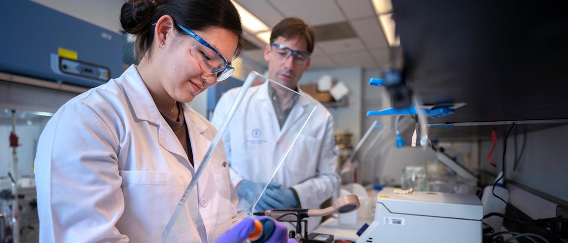 A woman in a white lab coat and goggles faces a counter full of laboratory equipment as a man in a white lab coat and goggles watches. 