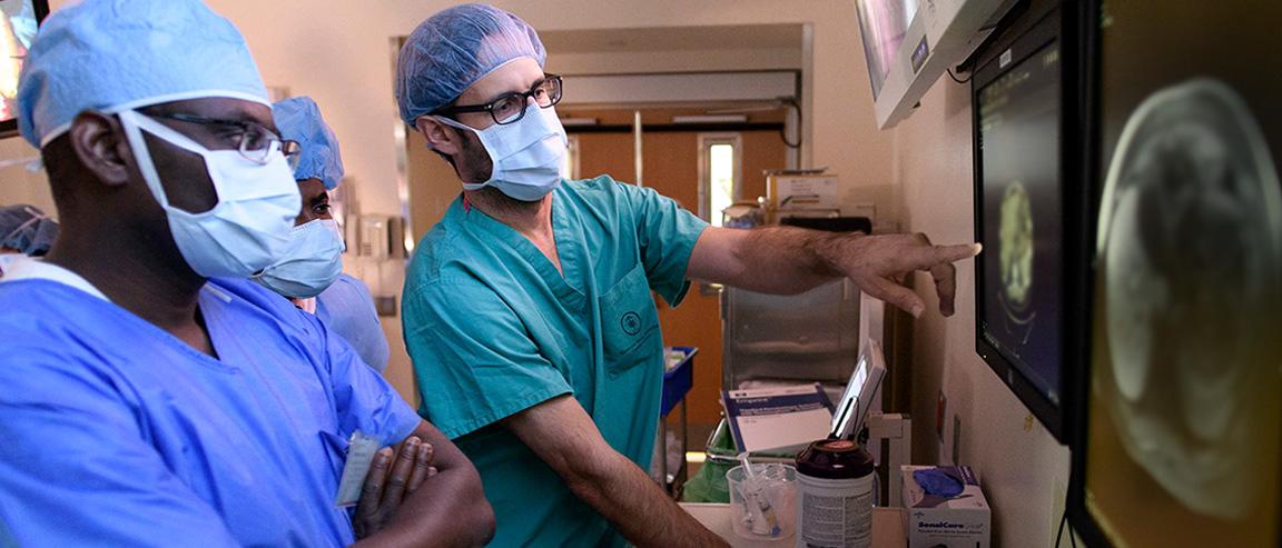 Three medical professionals wearing scrubs, face masks, and surgical caps look at an image on a screen.  