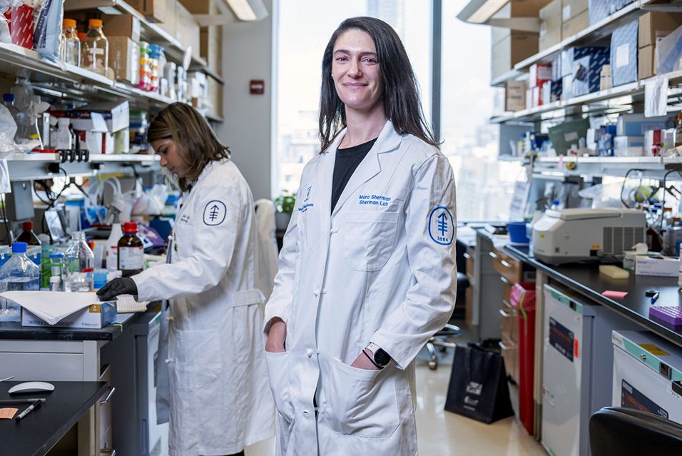 Scientist smiling in a busy laboratory, wearing a white lab coat.