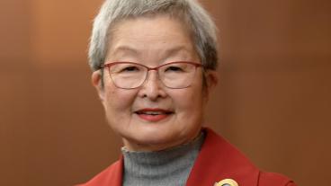 A woman with short gray hair in a gray mock turtleneck sweater and red blazer with a gold brooch stands, smiling, against a wood backdrop. 