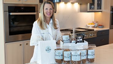 Granny-O-La founder Jennifer stands in a kitchen with a display of Granny-O-La products in front of her. 